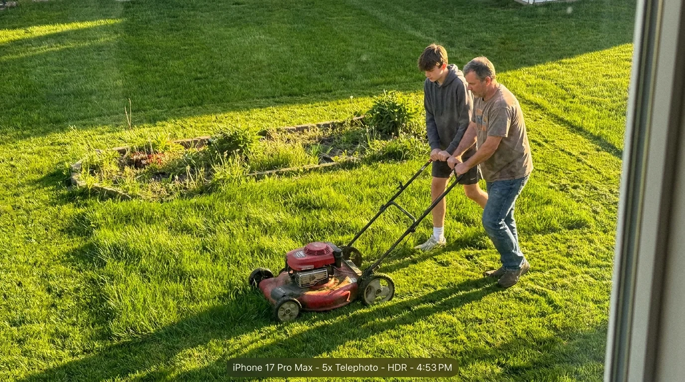 A high-angle photo of a parent and teenager mowing the lawn together in the golden light of late afternoon.