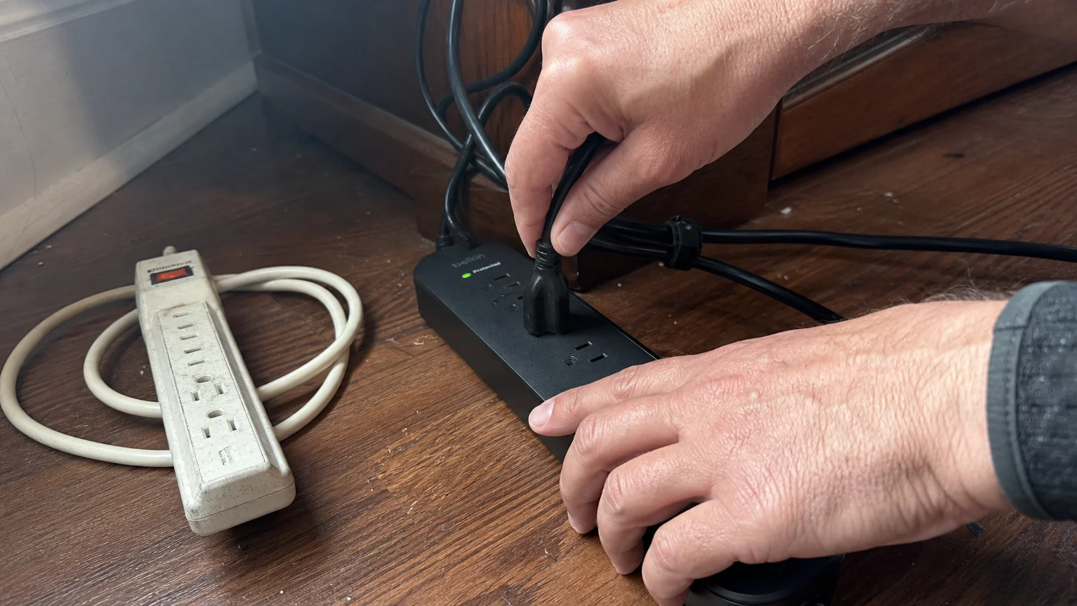 Hands plug a black cable into a new surge protector on a hardwood floor, next to a discarded yellowed power strip.