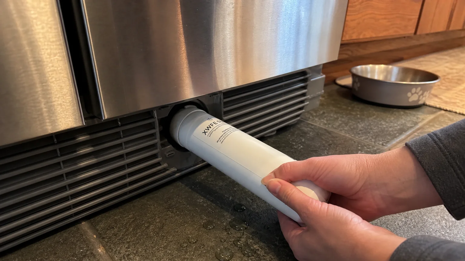 Hands twist a white cylindrical water filter out of the bottom compartment of a stainless steel refrigerator.