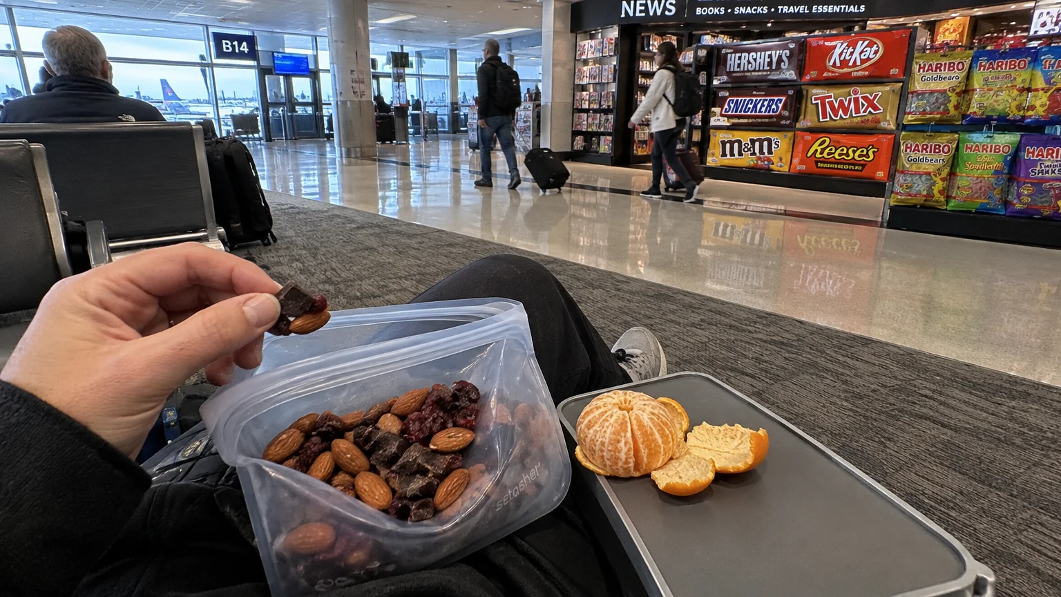 A traveler holds homemade trail mix from a reusable bag at an airport gate across from a candy-filled newsstand.