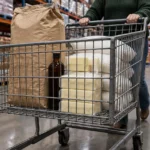 A shopper pushes a cart loaded with bulk flour, sugar bags, blocks of butter, and a bottle through a warehouse store.