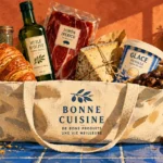Canvas tote bag filled with olive oil, jamón ibérico, a croissant, cheese, and vanilla ice cream on a blue tiled counter.